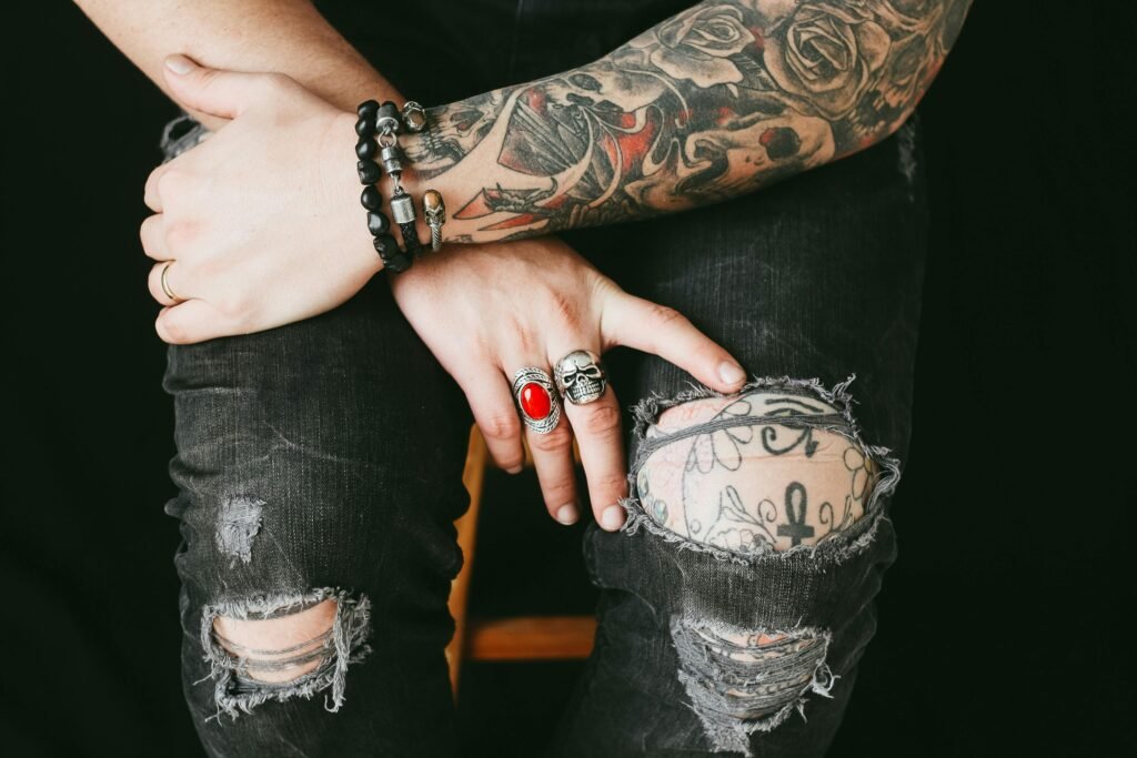 Close-up of tattooed hands with rings and bracelets over ripped jeans, showcasing fashion and style.
